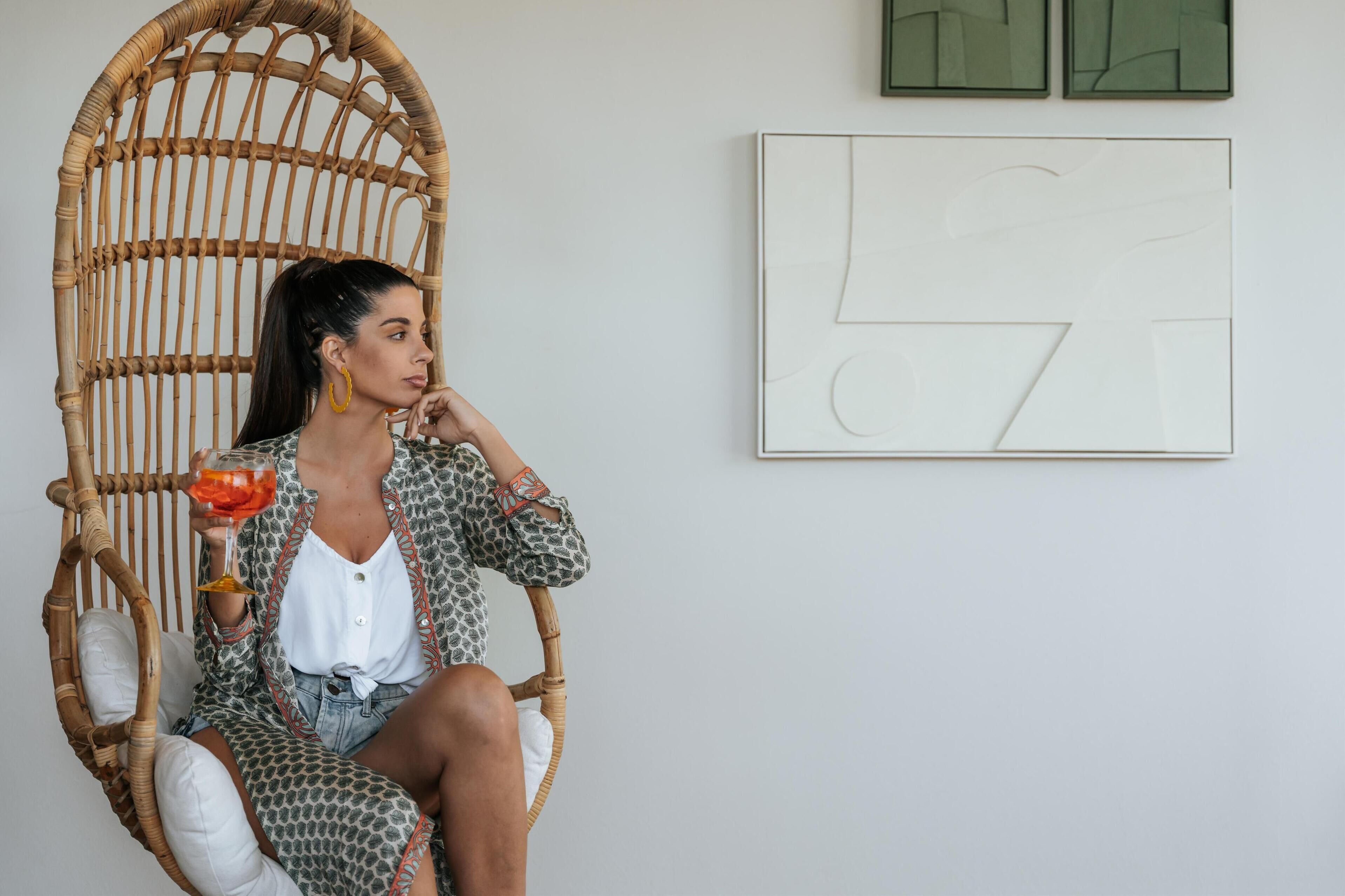 A person seated in a wicker chair with a white cushion, holding a drink.
