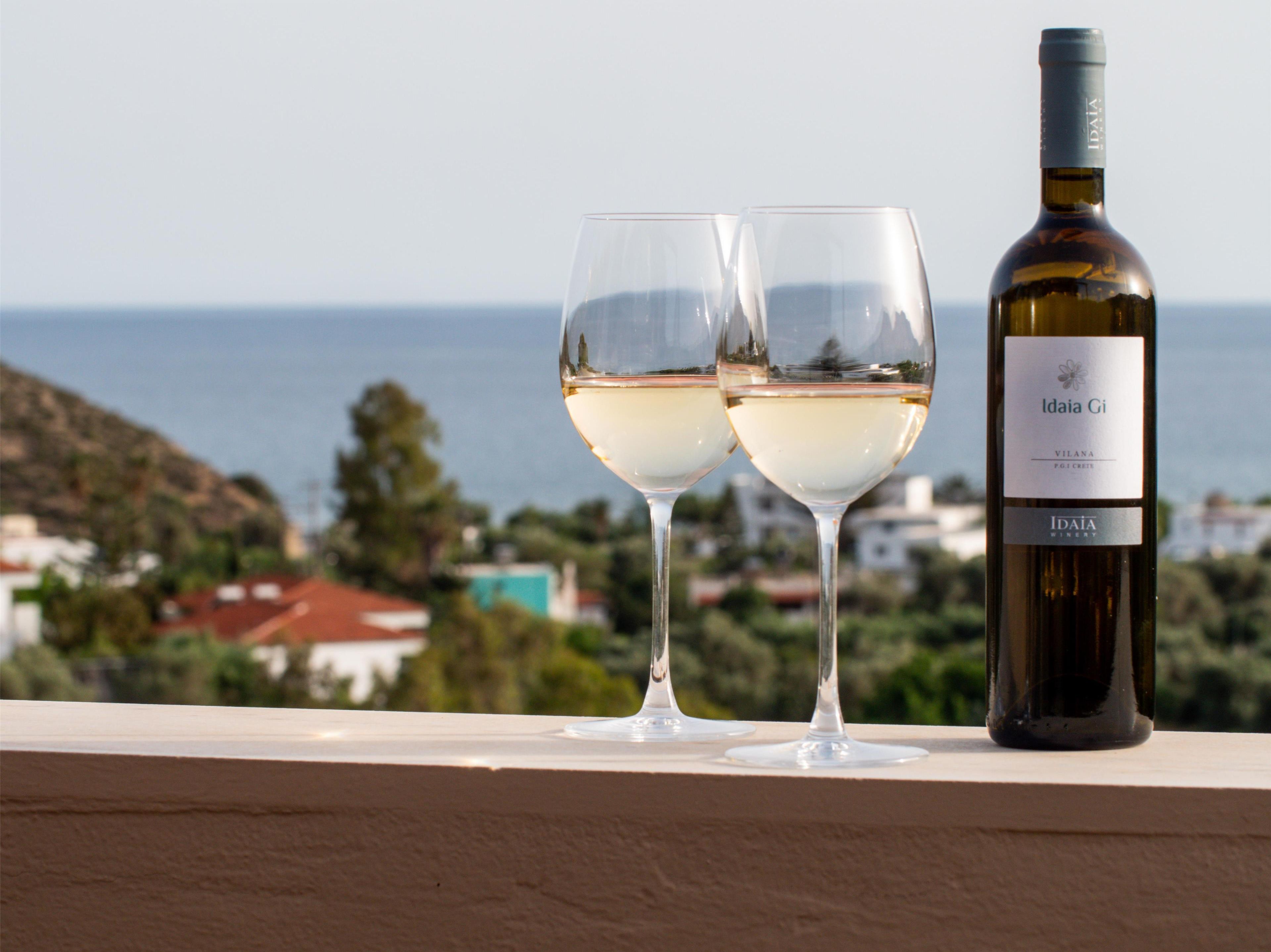 Eine Weinflasche und zwei Gläser auf einem Balkon mit Blick auf das Meer und Gebäude.