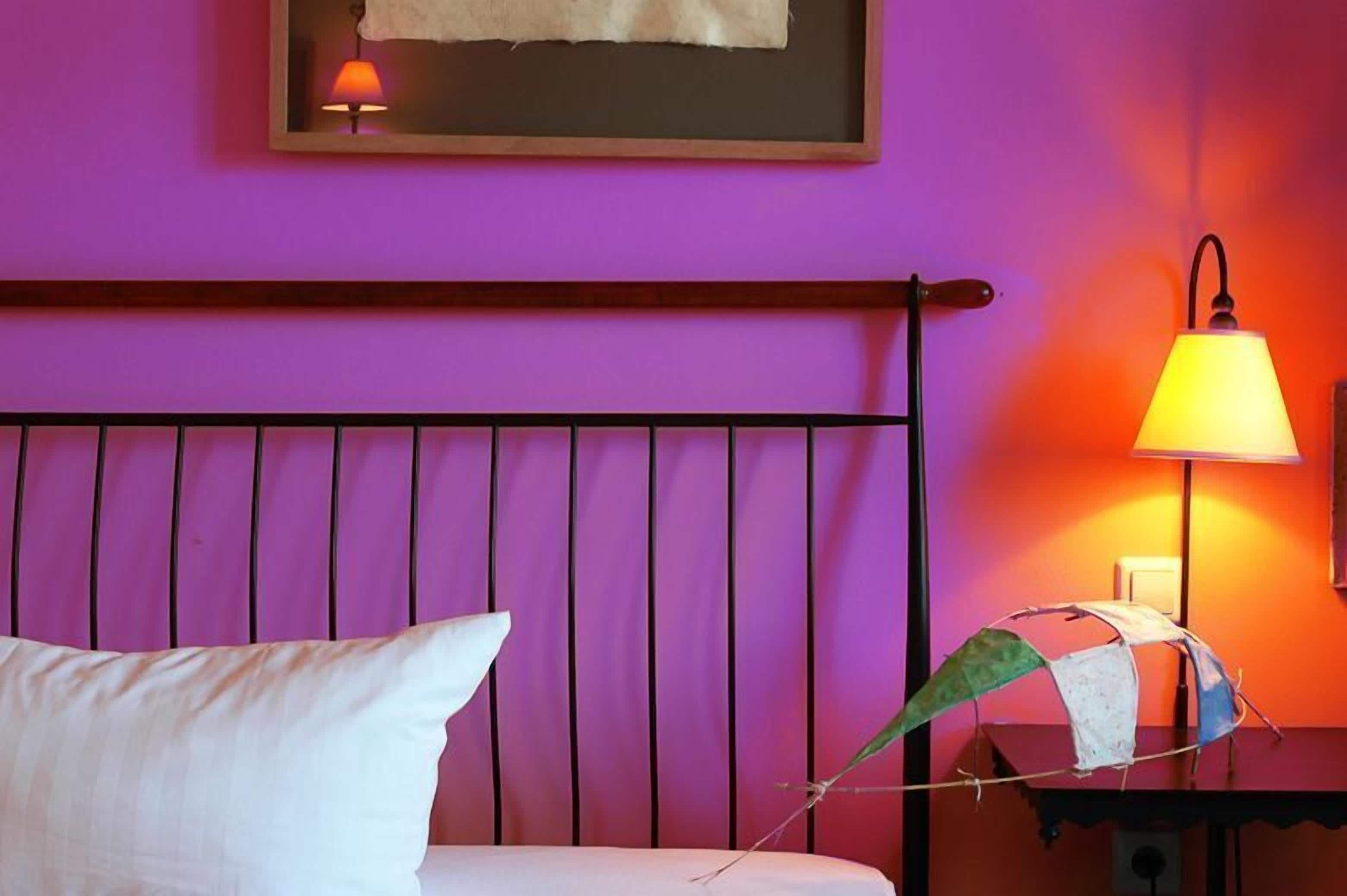 A bed with a white pillow and a metal headboard against a purple wall. A lamp with a yellow shade is on a bedside table.
