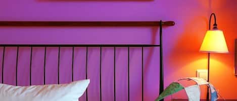 A bed with a white pillow and a metal headboard against a purple wall. A lamp with a yellow shade is on a bedside table.