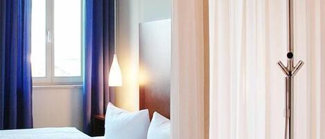 Habitación de hotel con una cama, una ventana con cortinas azules, una lámpara de noche y zona de ducha con cortina.