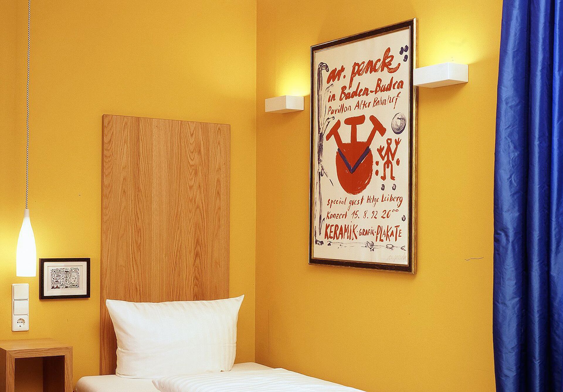 Habitación de hotel con una cama, cabecero de madera, un cartel en la pared, una lámpara y una cortina azul.