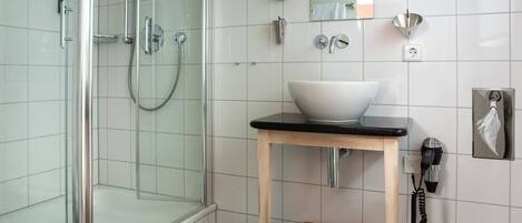 Un baño moderno con cabina de ducha de vidrio, un lavamanos blanco sobre encimera negra y una cesta de mimbre.