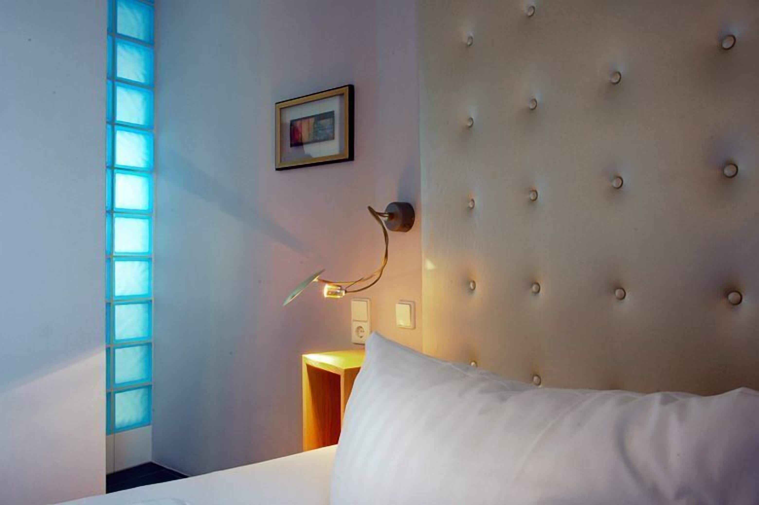 A hotel room with a tufted headboard, a bedside lamp, a framed picture on the wall, and a window with blue-tinted glass panels.