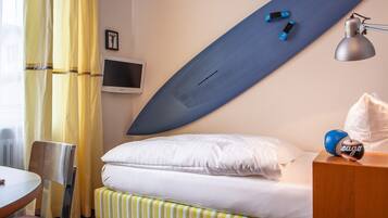 A hotel room with a bed, a desk, a chair, a TV, and a surfboard on the wall.