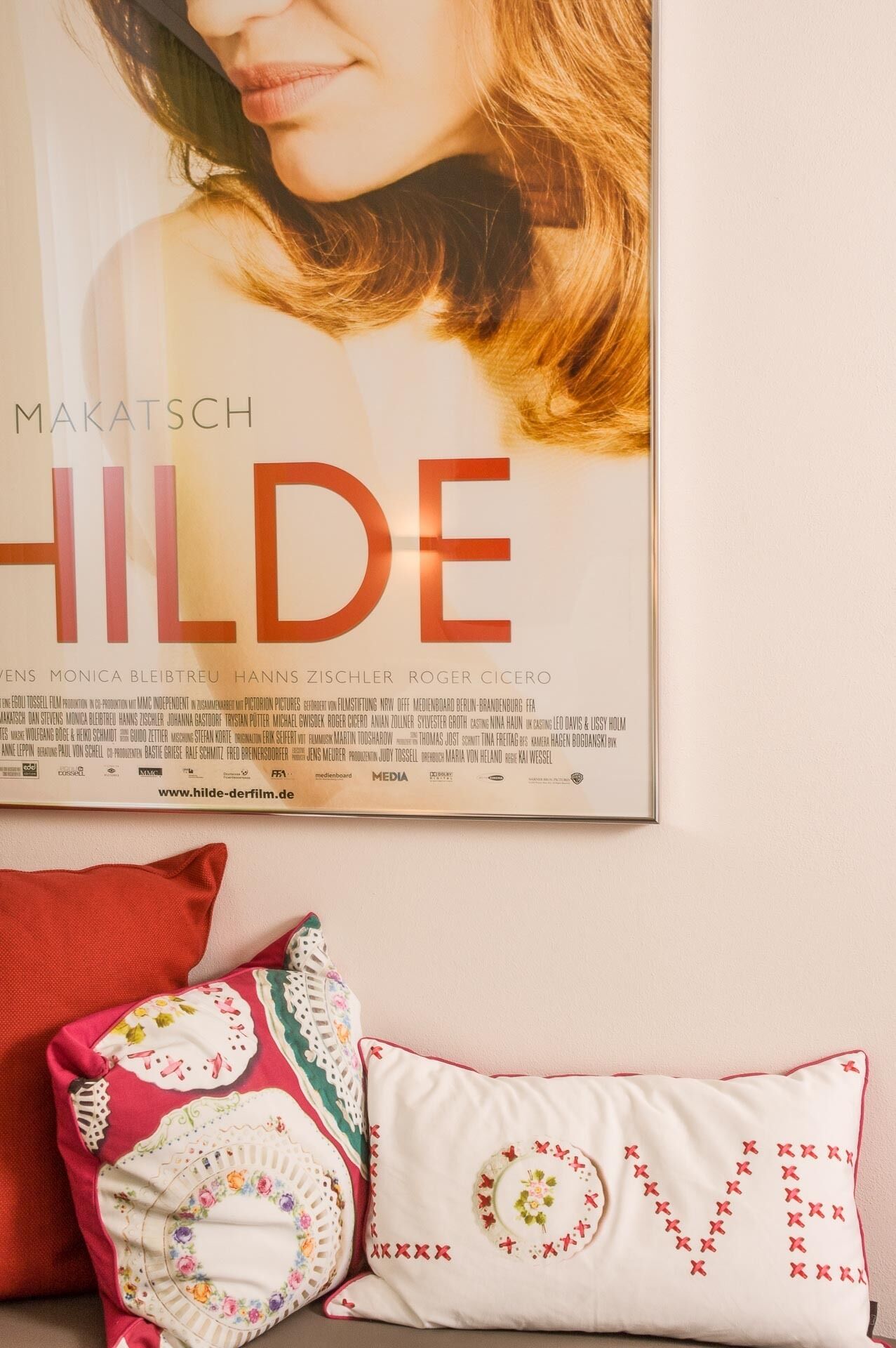 Un cartel de película enmarcado de 'MAKATSCH HILDE' en una pared, con dos almohadones decorativos debajo.