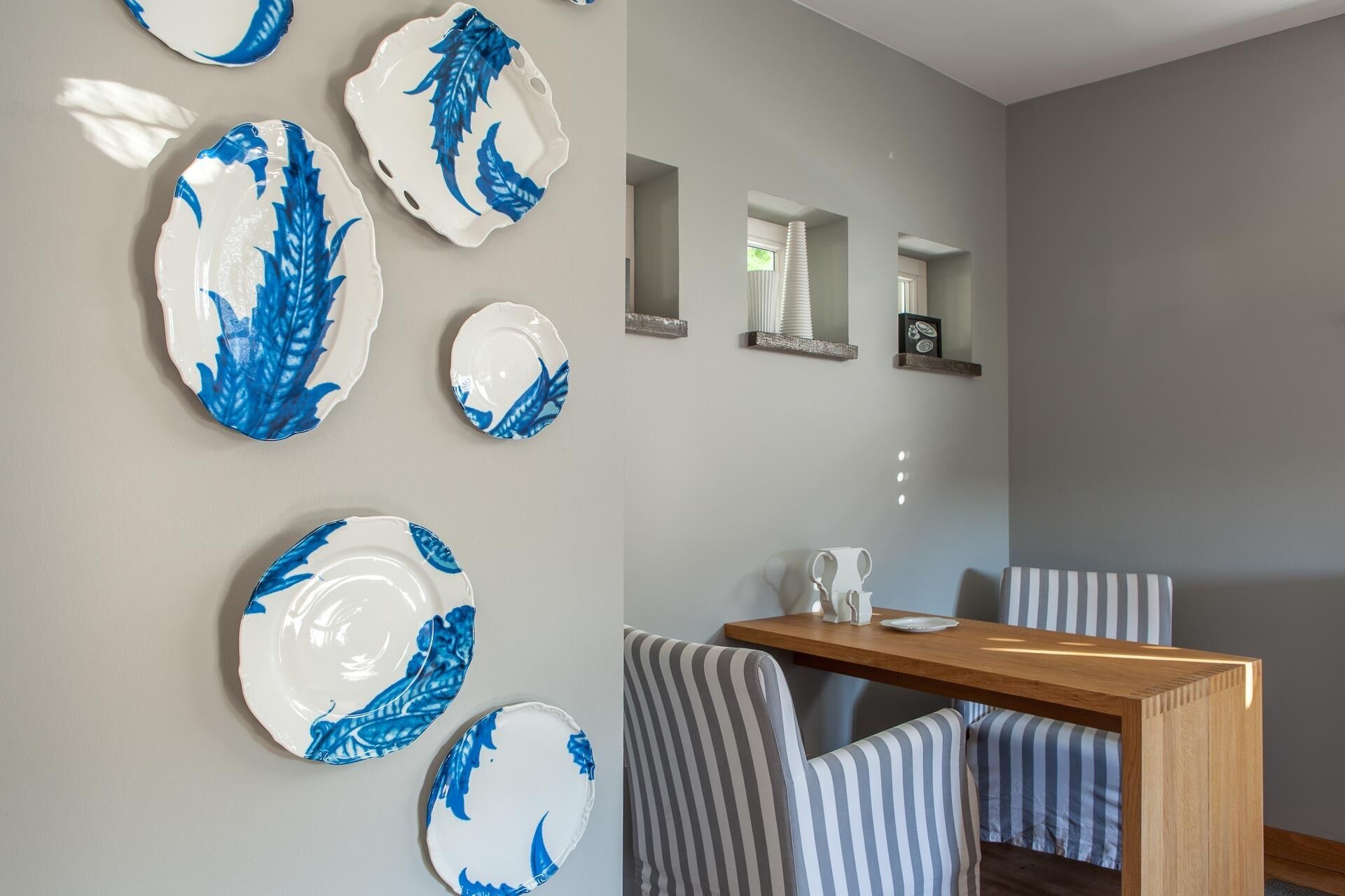 Un comedor con una mesa de madera y sillas a rayas, una pared decorada con platos, y un espejo sobre una repisa.