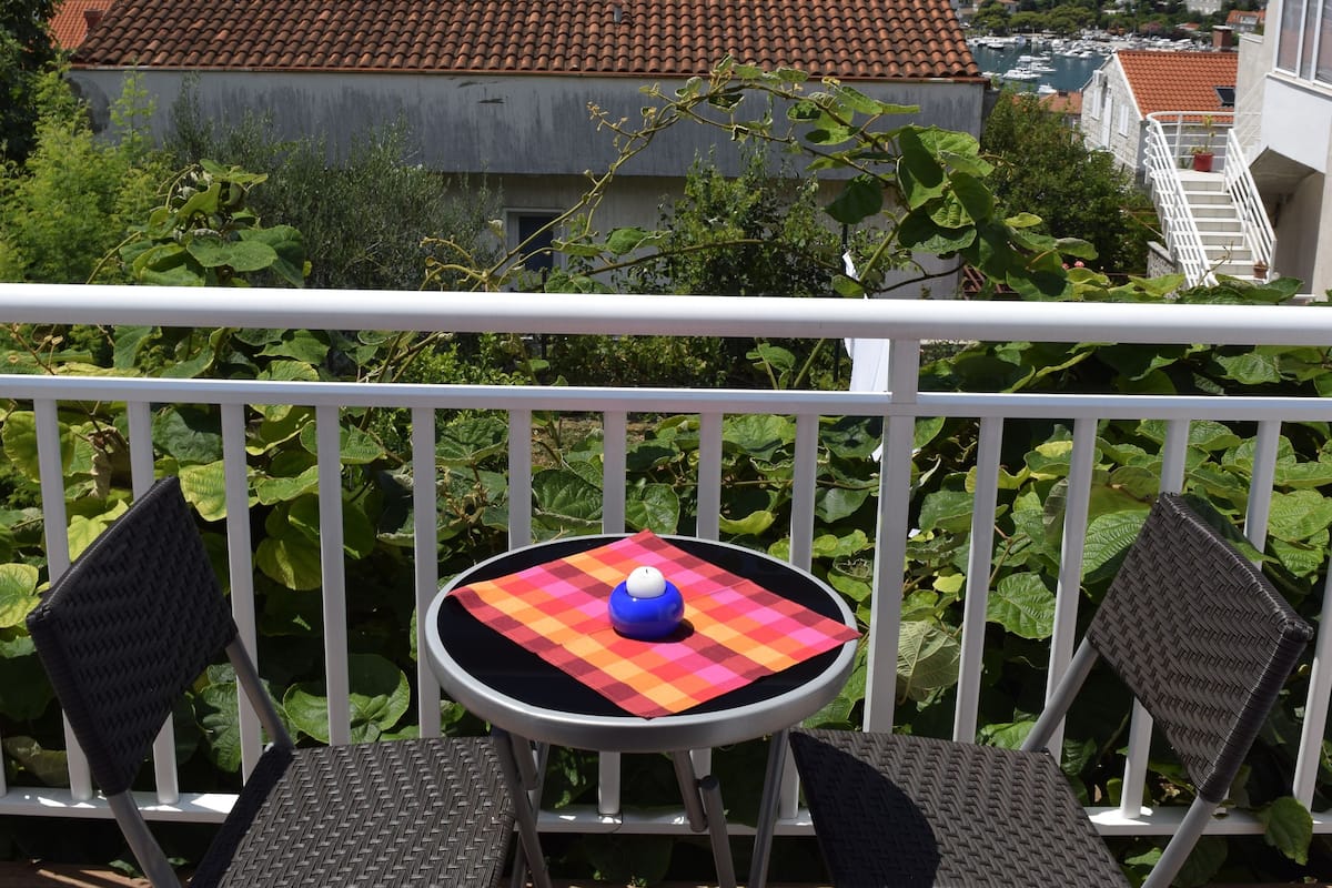 A balcony with two chairs and a small table, overlooking a hillside with terracotta-roofed buildings and greenery.