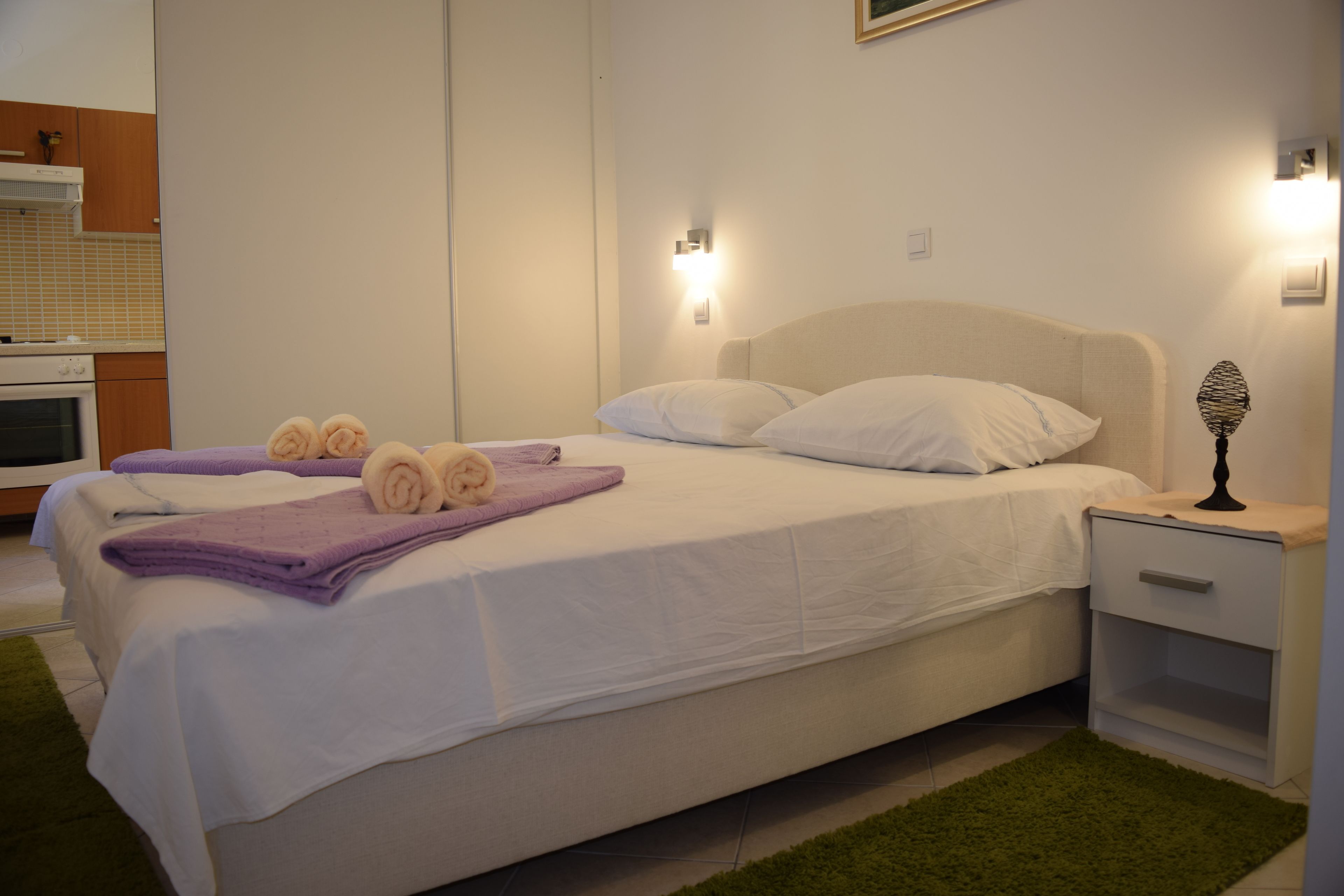 A neatly made bed with white linens and rolled towels, a bedside table with a lamp, and a green rug.