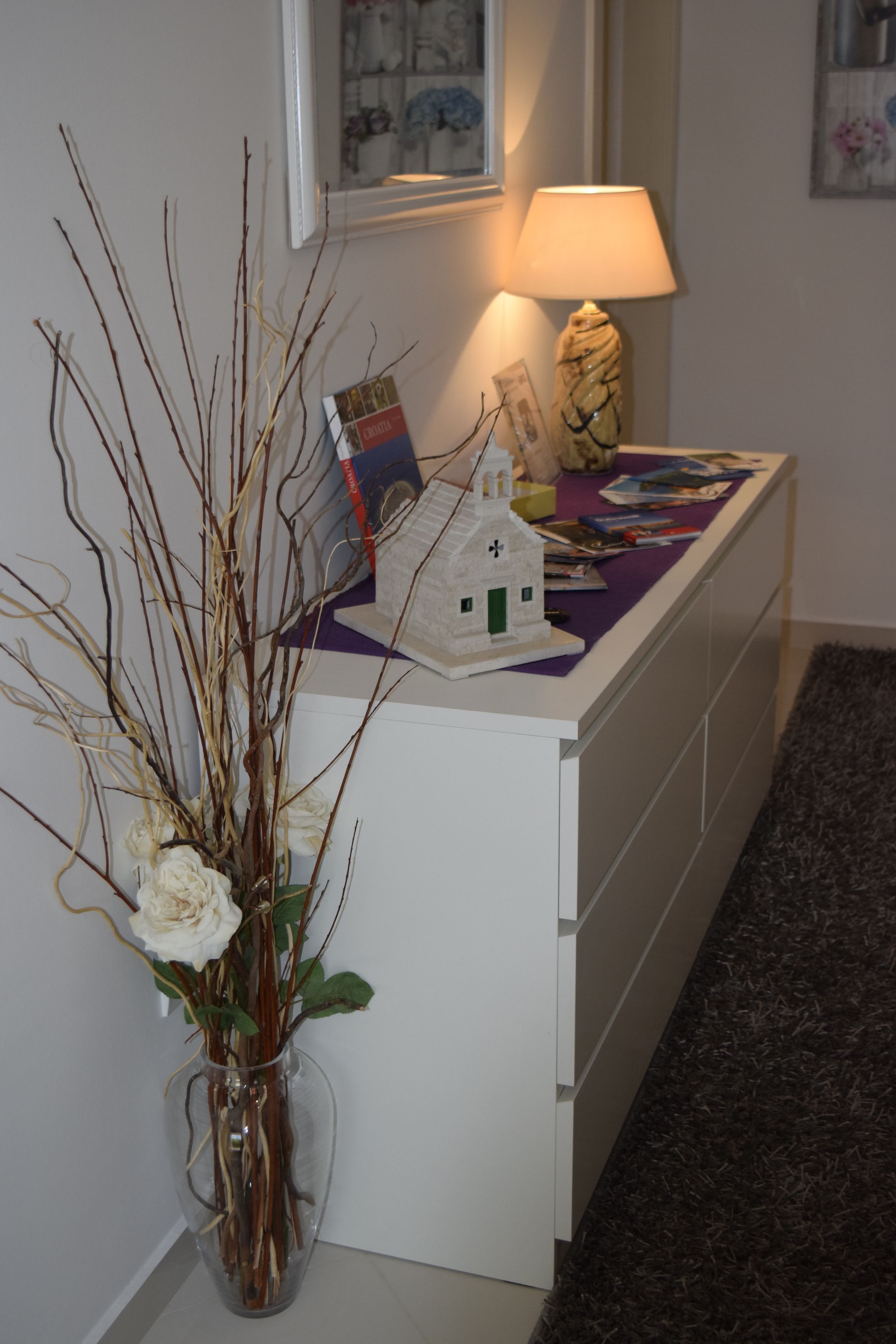 Un bureau blanc orné d’un vase contenant des branches séchées et des fleurs blanches, une petite maquette de maison blanche et une lampe posée sur un tissu violet.
