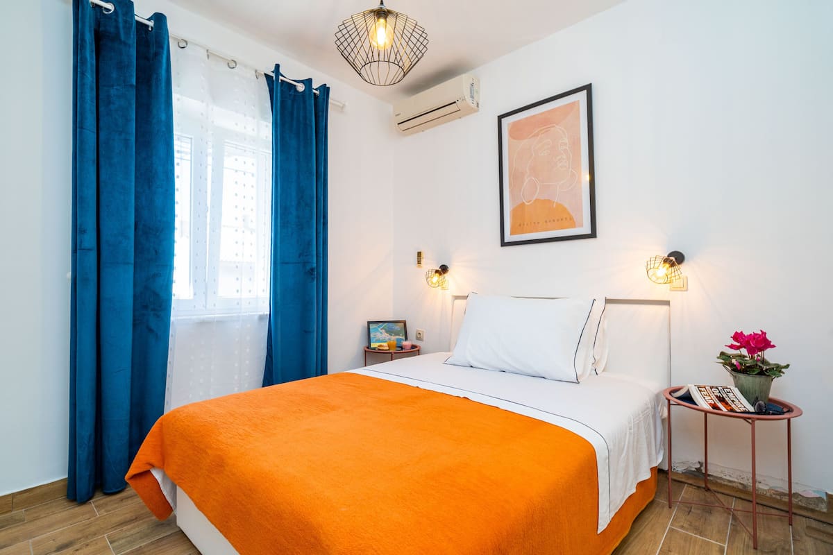 A bedroom with a bed, curtains, a wall-mounted air conditioner, a framed picture, bedside lamps, a small table with a magazine and a flower pot.