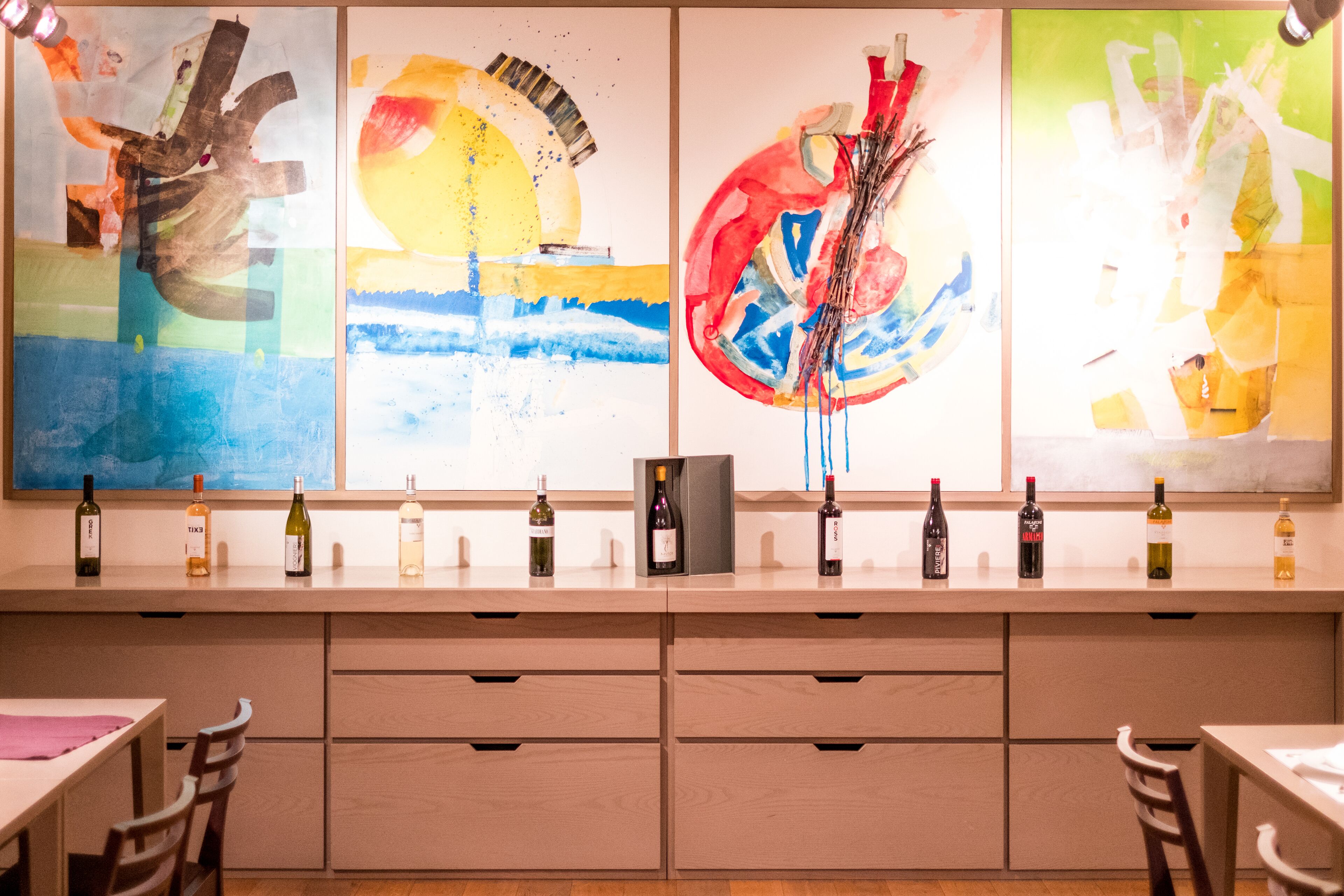 Un espace bar moderne avec un long comptoir où sont exposées différentes bouteilles, une grande peinture abstraite au mur et un aménagement intérieur minimaliste.