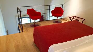 Une pièce avec un plancher en bois, un lit recouvert d’une couverture rouge et deux chaises rouges.
