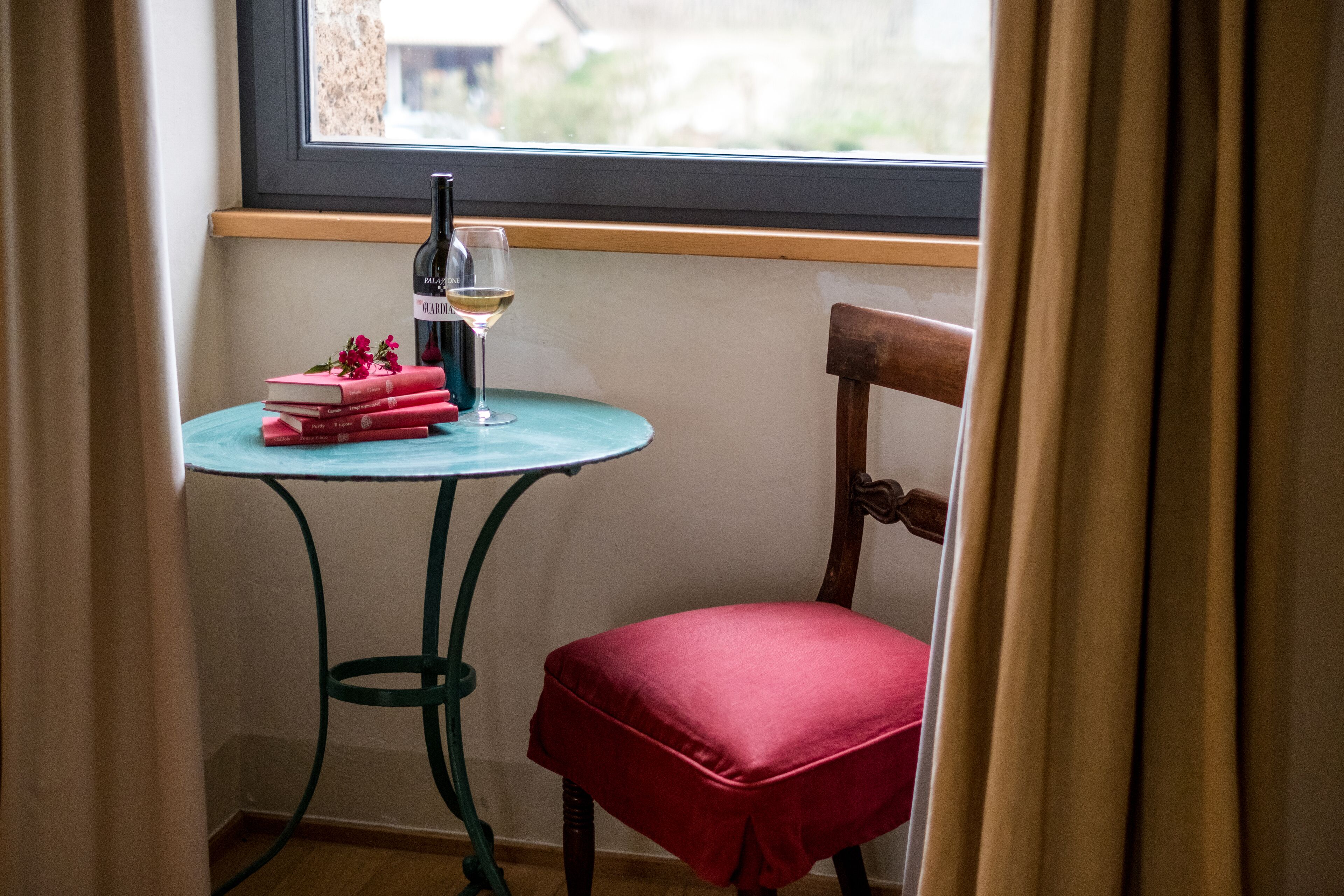 Una habitación de esquina con ventana, una pequeña mesa redonda con una botella de vino y libros, y un sillón rojo acolchado.