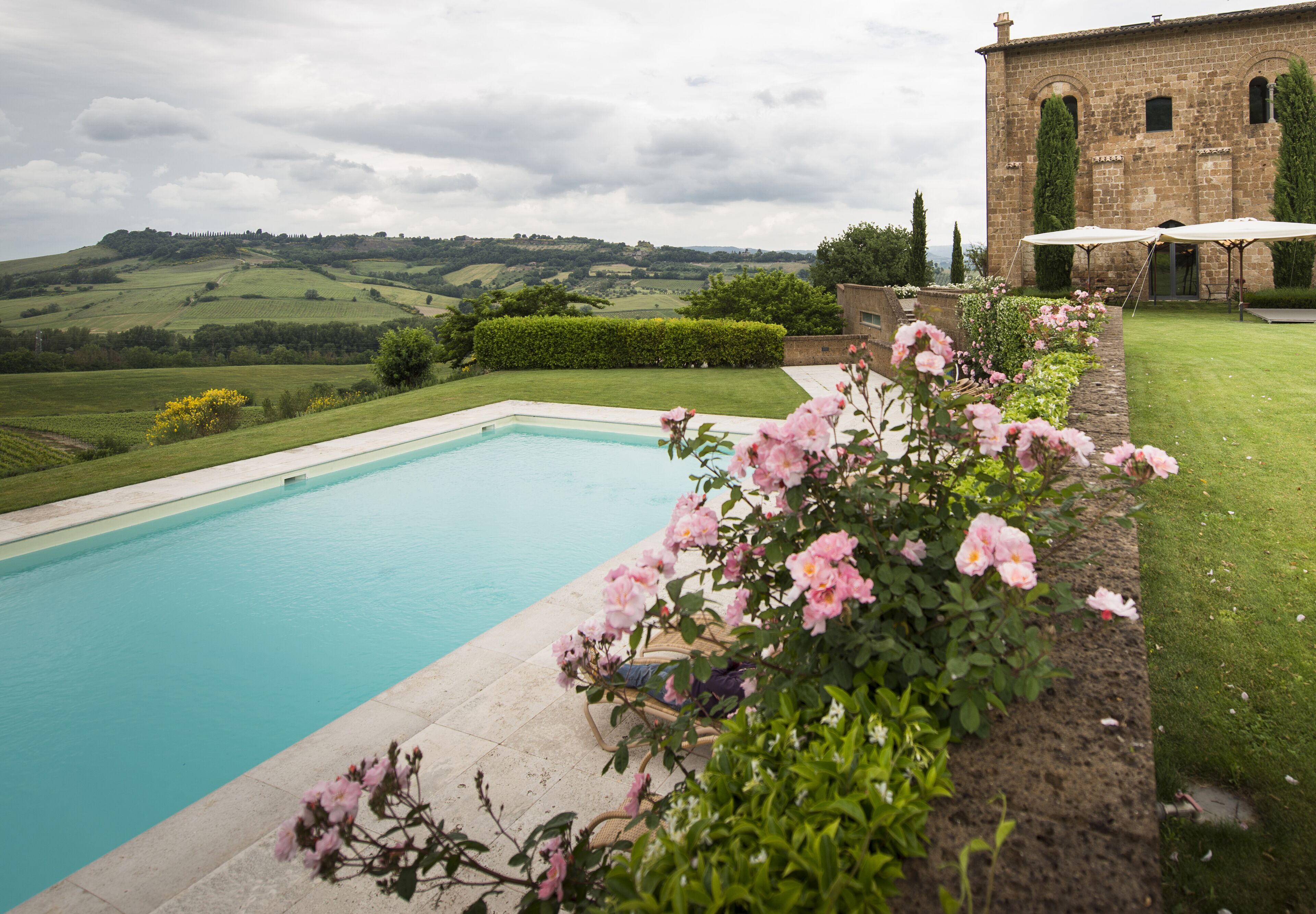 A swimming pool with a view of rolling hills, a brick building, and a garden with pink flowers.