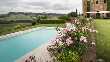 A swimming pool with a view of rolling hills, a brick building, and a garden with pink flowers.