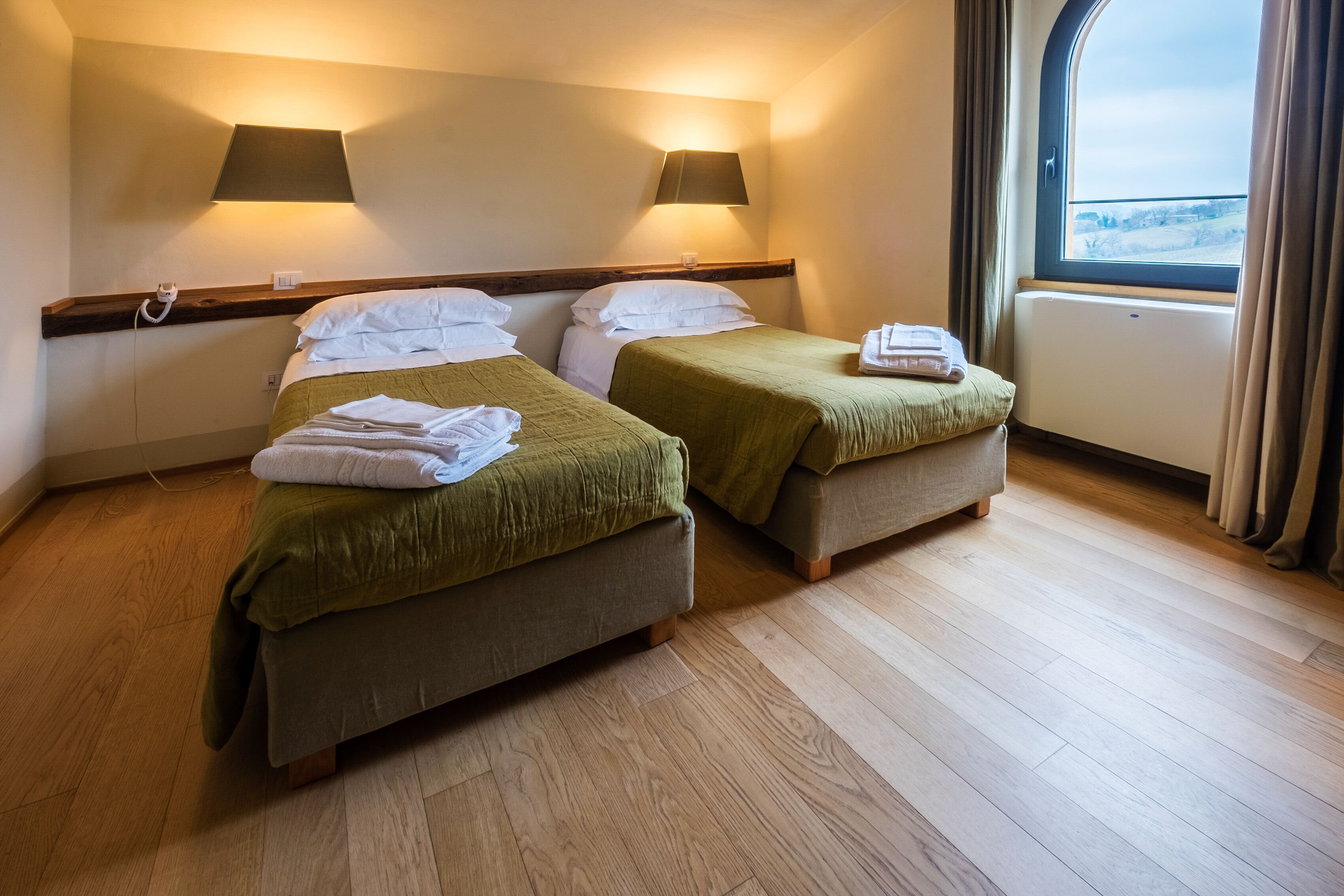Quarto com duas camas, cada uma com colcha verde e almofadas brancas, chão de madeira e janela com cortinas.
