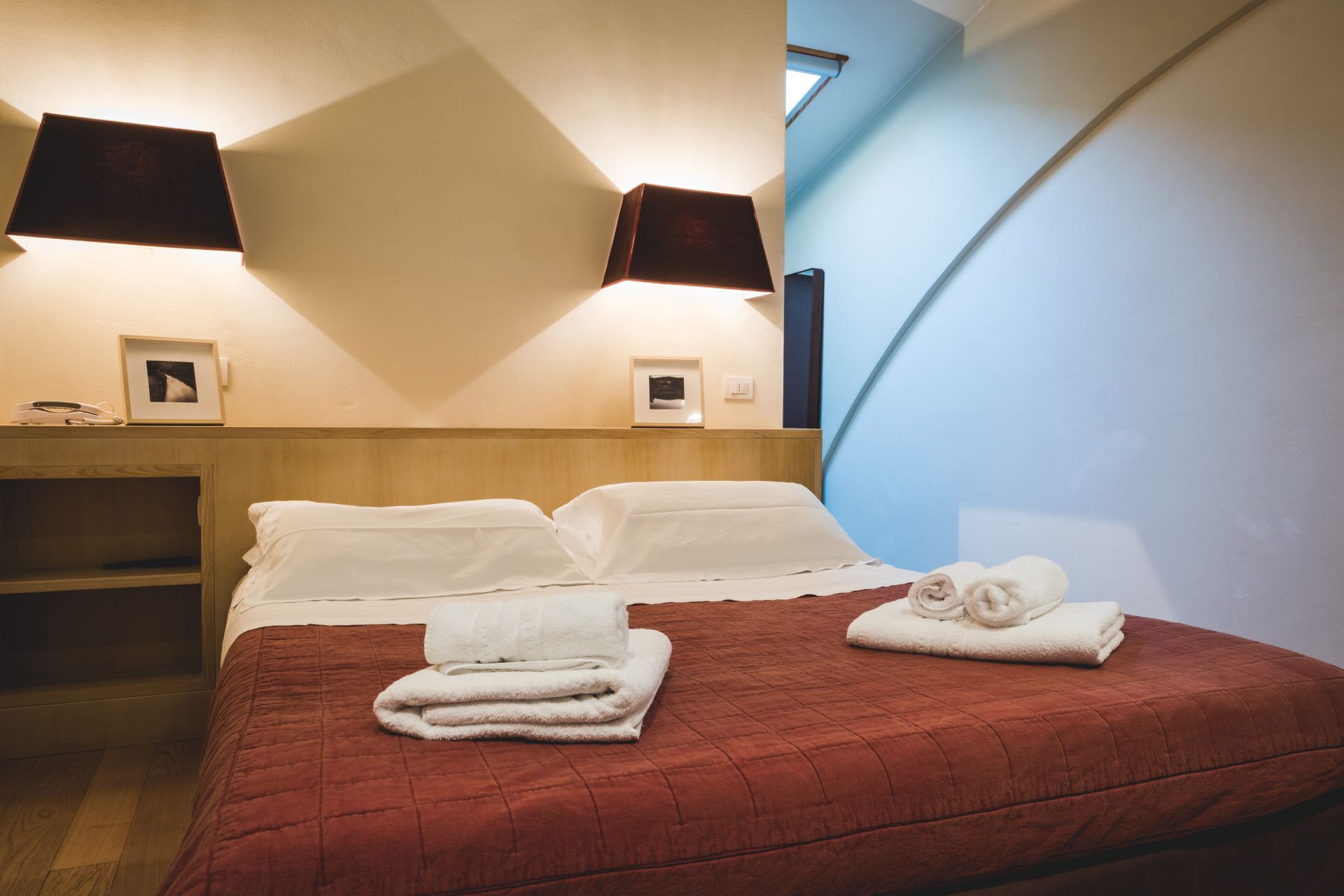 Habitación de hotel con cama, dos lámparas adosadas a la pared, cabecero de madera y ventana con vistas.