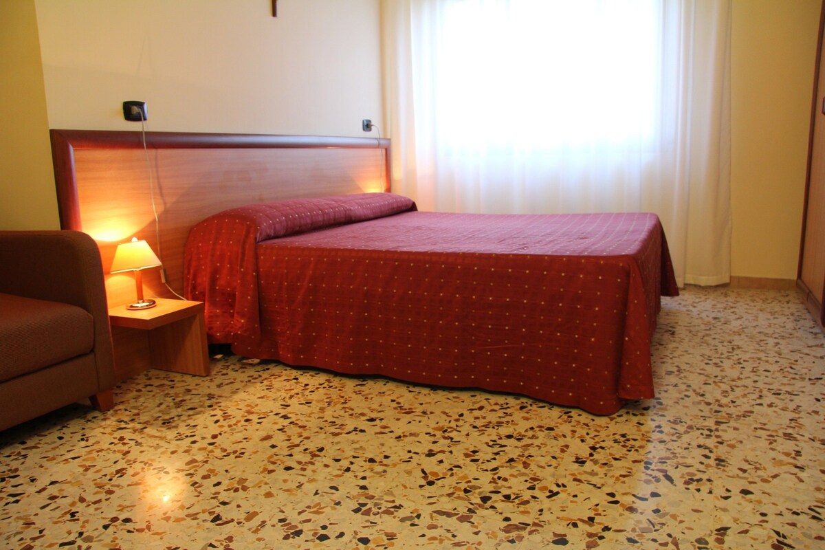 Une chambre d’hôtel avec un lit, une table de chevet avec une lampe, une chaise et une fenêtre avec des rideaux.