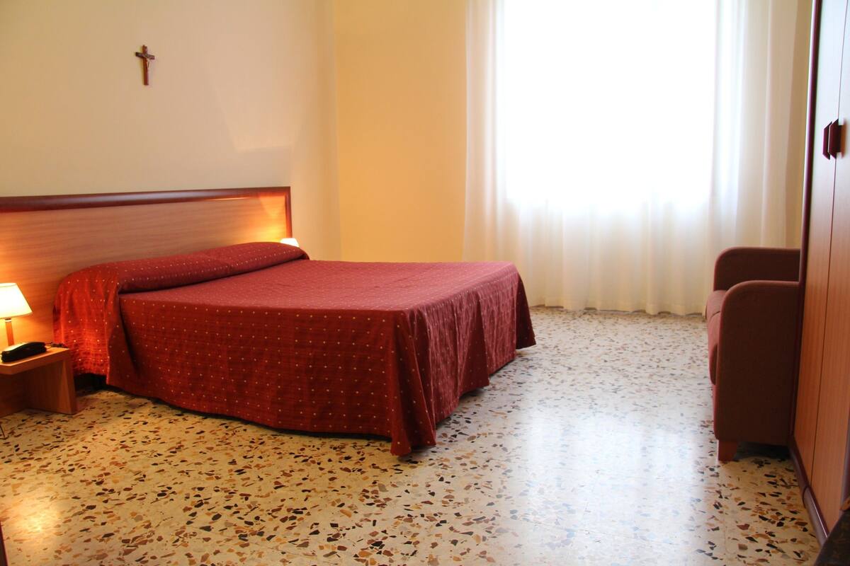 Une chambre d’hôtel avec un lit, une table de chevet, une chaise et une croix accrochée au mur.