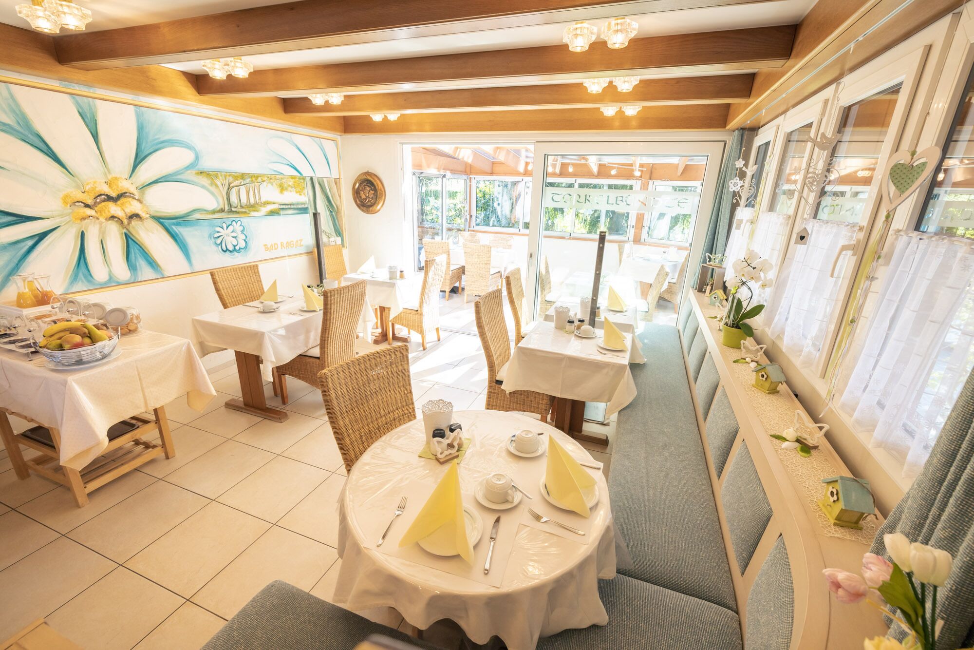 Een restaurant met ronde tafels, rieten stoelen en een muurschildering van een bloem.