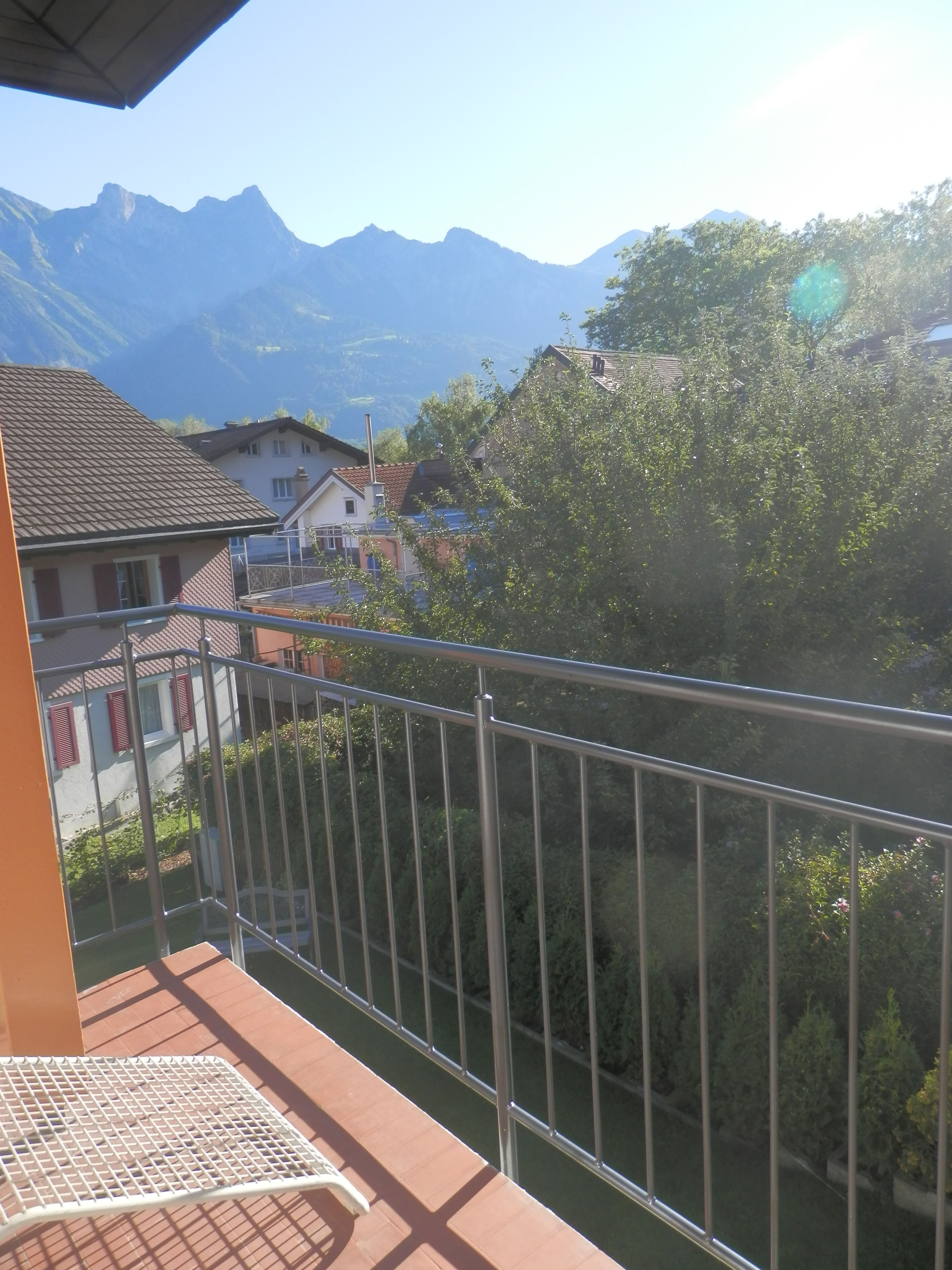 A balcony with a metal railing overlooking a mountainous landscape and residential buildings.