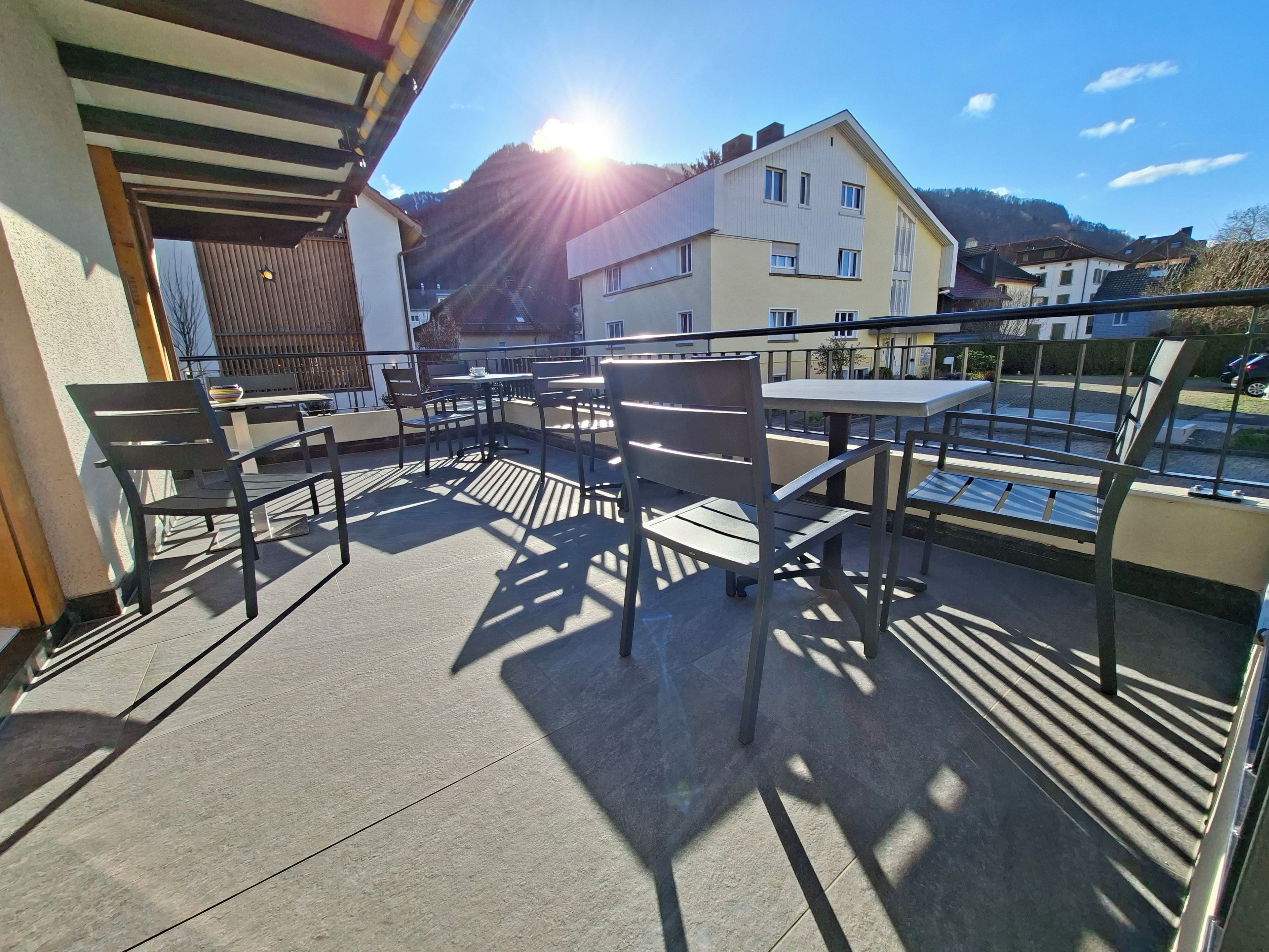 A balcony with outdoor seating, a view of buildings, and a mountain in the background.
