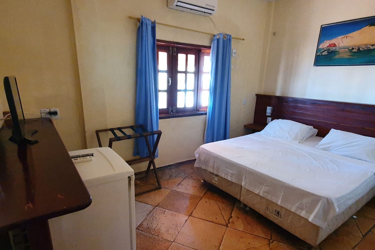 A room with a bed, a desk, and a chair. There is a window with curtains, an air conditioning unit, and a painting on the wall.