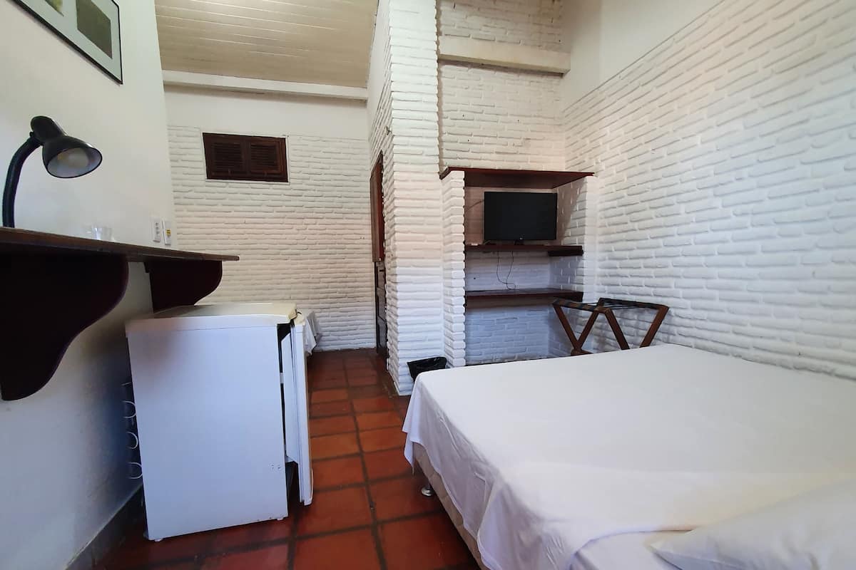 A compact room with a bed, a small desk, a white refrigerator, a TV mounted on a brick wall, and a wall-mounted shelf.
