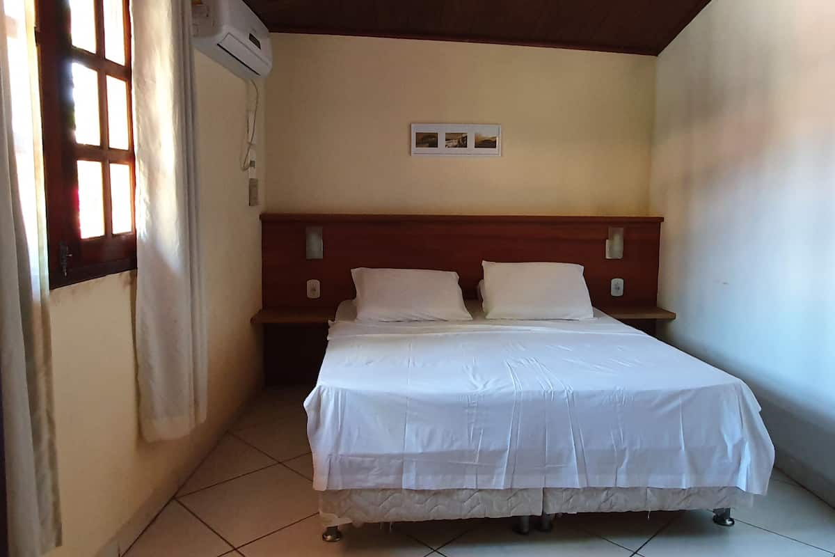A hotel room with a bed, a wooden headboard, a wall-mounted air conditioning unit, a small bedside table, and a window with curtains.