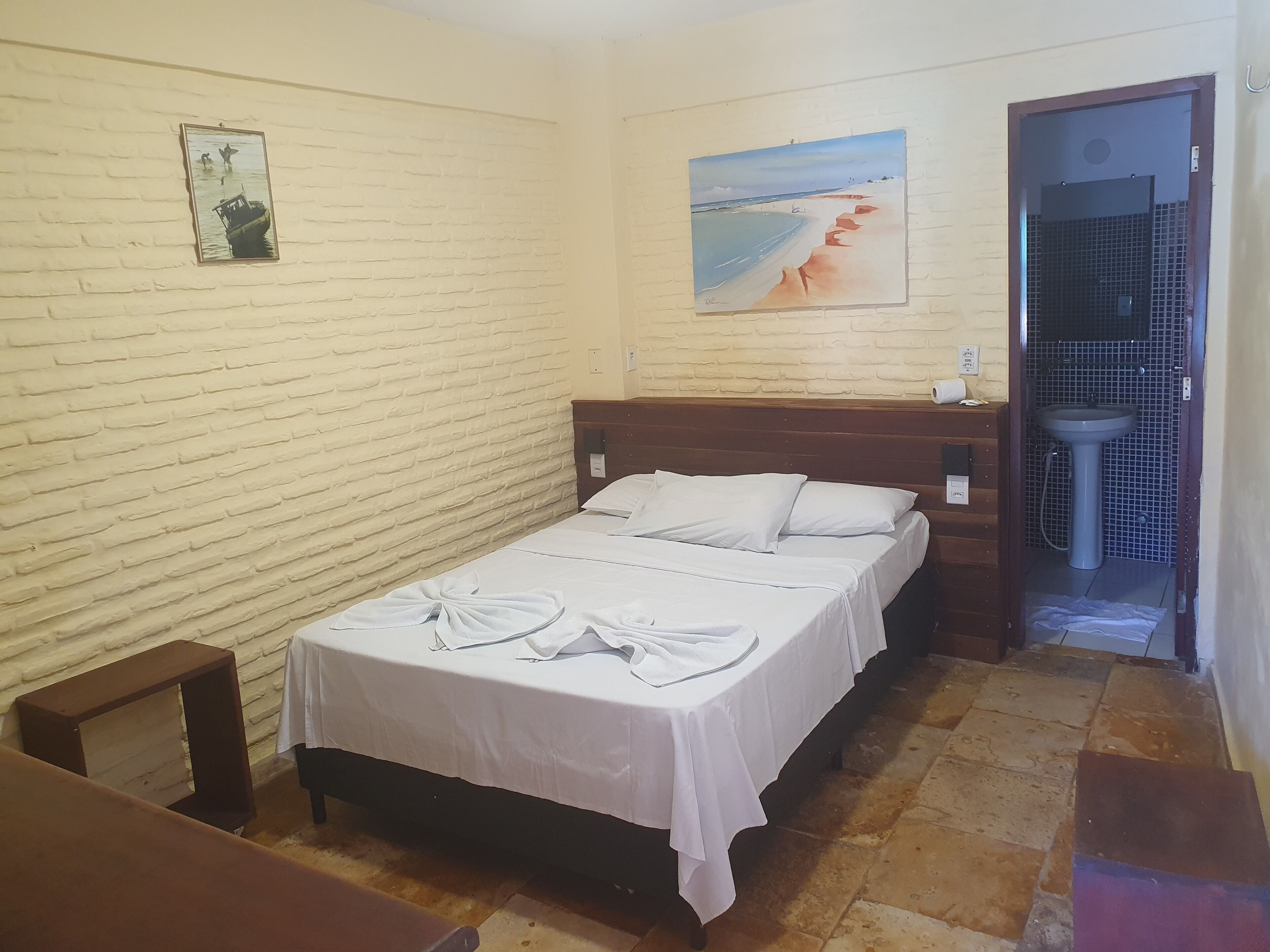 A hotel room with a brick-patterned wall, a bed with white linens, a painting of a beach, and a bathroom with a sink and tiled walls.