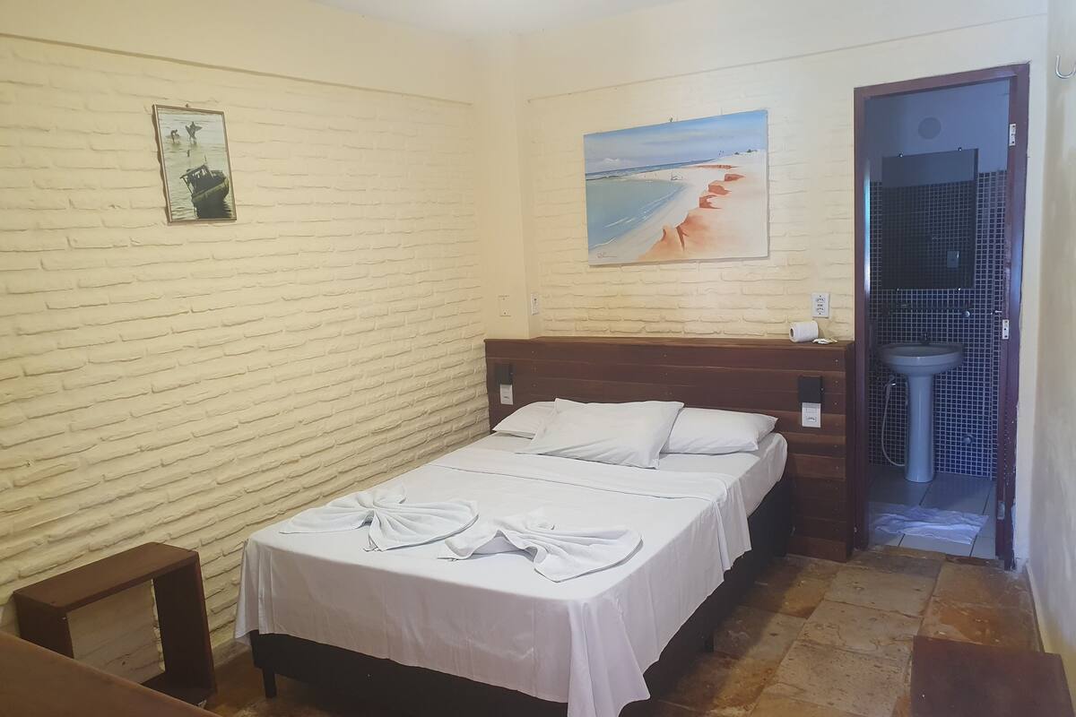 Une chambre d’hôtel avec un mur décoré d’un motif en briques, un lit avec du linge de maison blanc, un tableau représentant une plage et une salle de bain avec un lavabo et des murs carrelés.