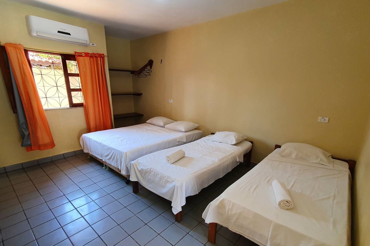 Une chambre avec deux lits simples, un appareil de climatisation, une fenêtre avec des rideaux orange et un sol carrelé.