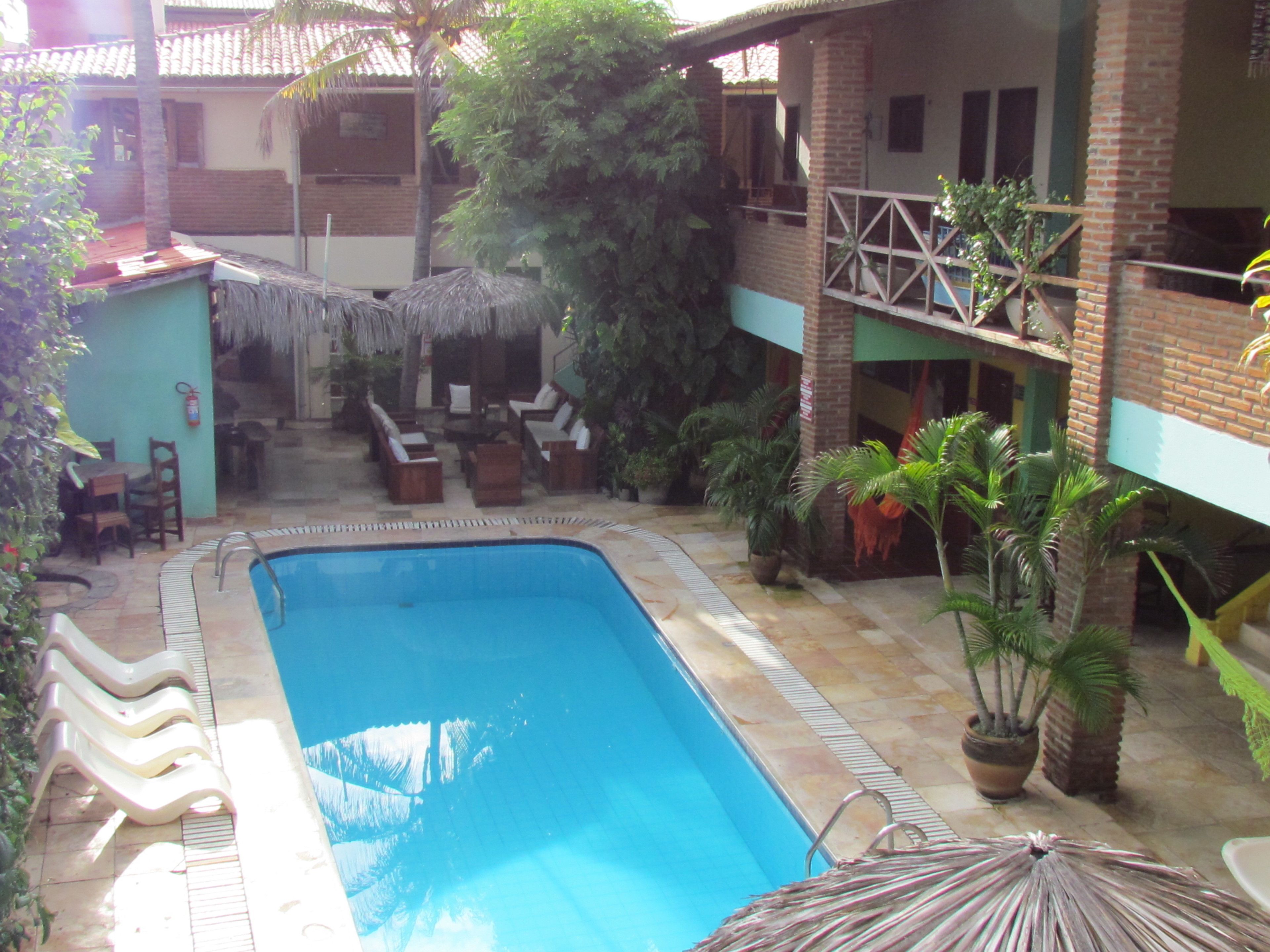 Piscina com espreguiçadeiras e guarda-sóis, cercada por vasos de plantas e um edifício de tijolos.