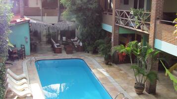 Piscina com espreguiçadeiras e guarda-sóis, cercada por vasos de plantas e um edifício de tijolos.
