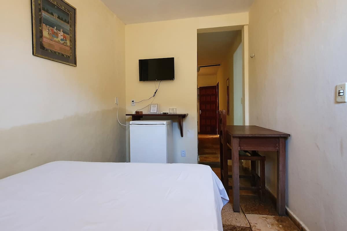 A small, narrow room with a bed, a desk, a television mounted on the wall, and a refrigerator.