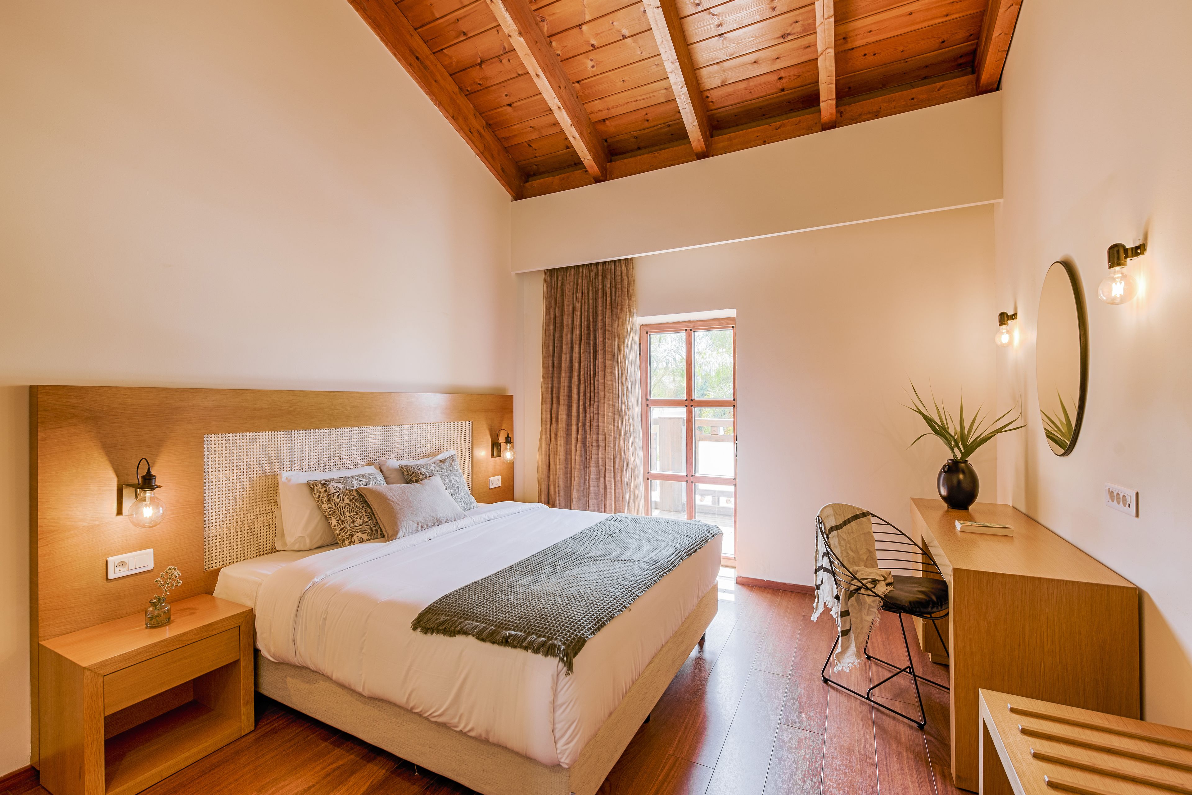 Een slaapkamer met een houten plafond, een groot bed met hoofdeinde, bedlampen, een bureau met stoel, en een raam met gordijnen.