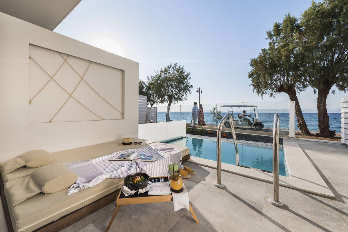 Un espacio exterior moderno con piscina, sofá, mesa de comedor con comida y vista al mar.