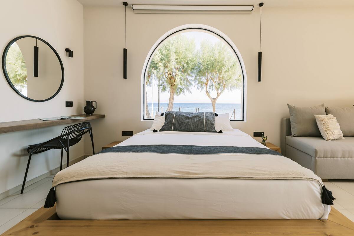 Un dormitorio con una cama grande, un escritorio con silla, un sofá y una ventana con vistas a árboles y al mar.