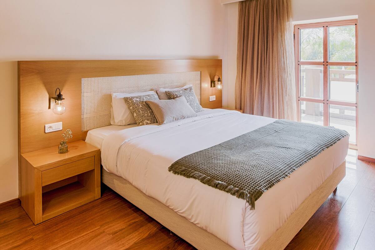 Un dormitorio con techo de madera, una cama grande con ropa blanca, una mesita de noche y una ventana con cortinas.