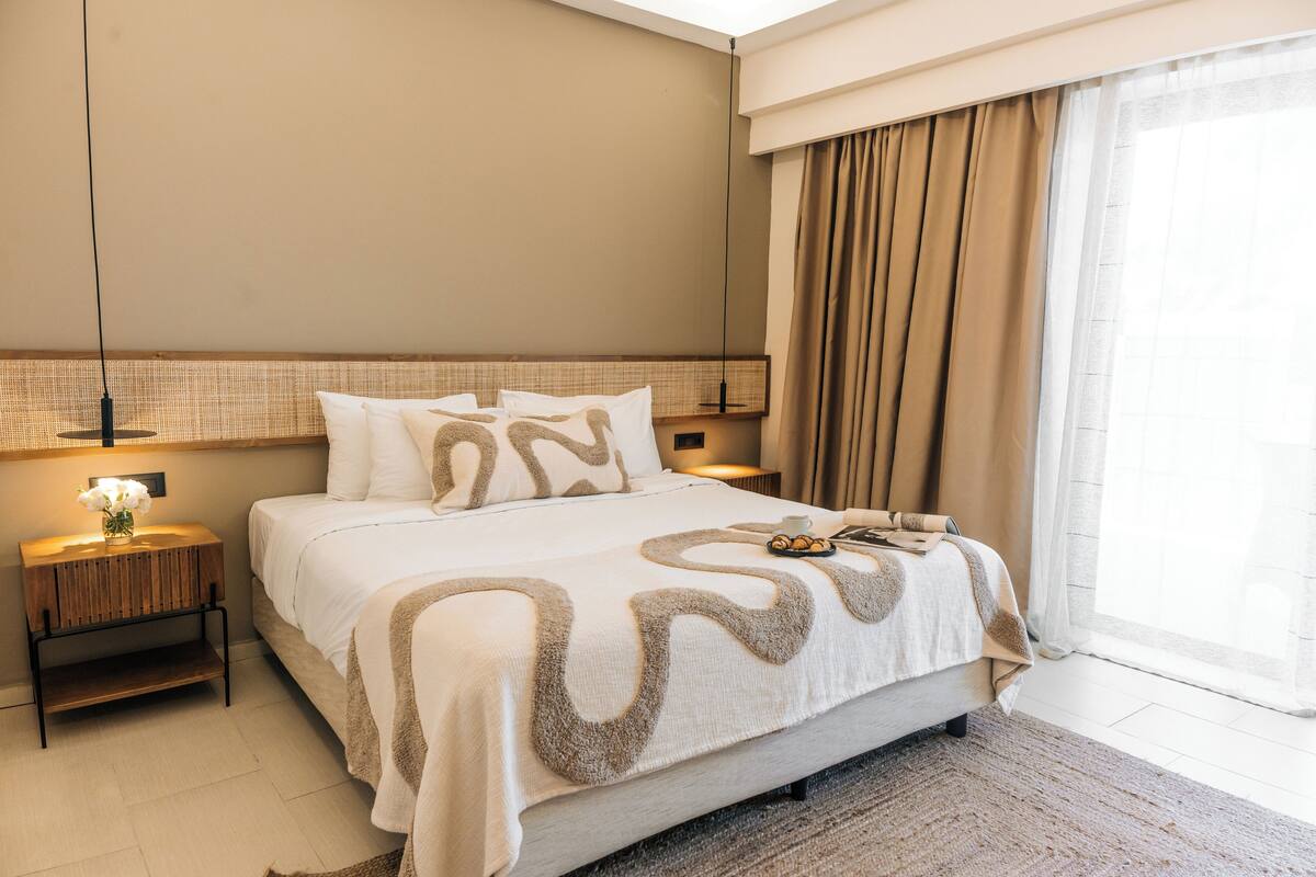 Una habitación de hotel moderna con una cama grande, una mesita de noche con lámpara y vistas al exterior a través de un ventanal.