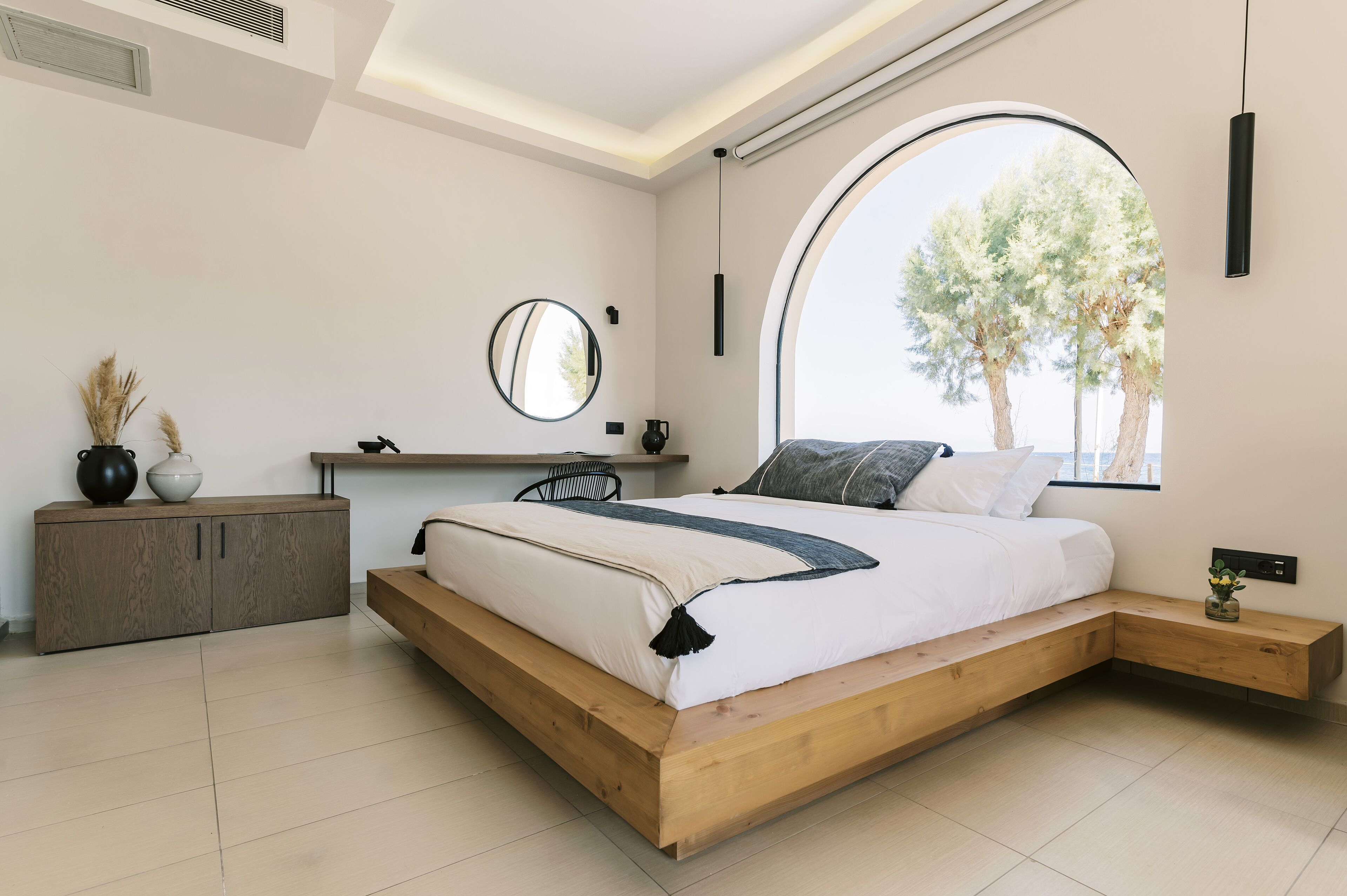 Ein modernes Schlafzimmer mit einem hölzernen Bett, einem runden Spiegel, einem Nachttisch und einem großen Fenster mit Blick auf Bäume.