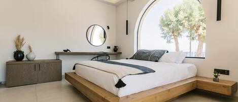 Ein modernes Schlafzimmer mit einem hölzernen Bett, einem runden Spiegel, einem Nachttisch und einem großen Fenster mit Blick auf Bäume.