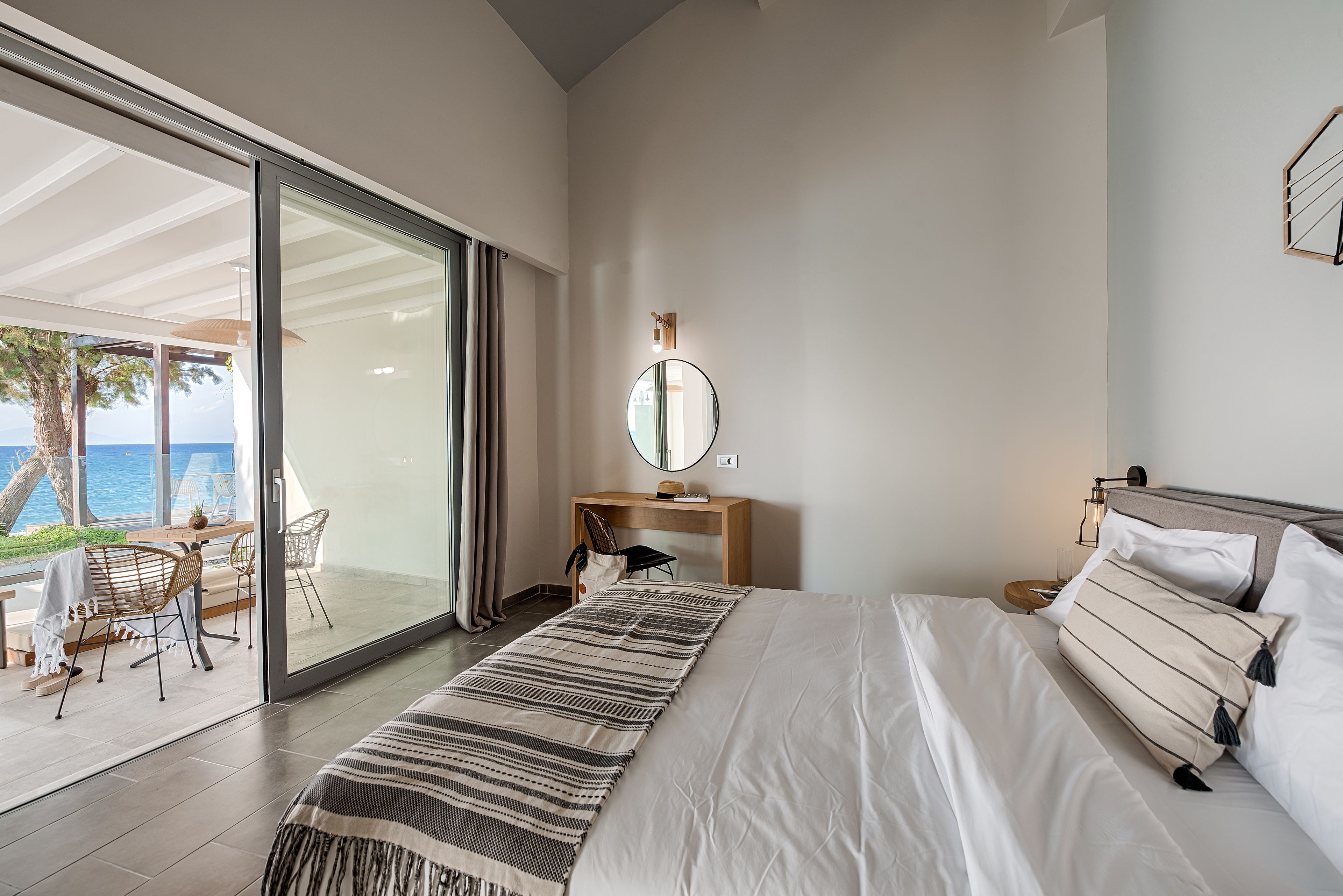 A bedroom with a bed, a desk, a chair, and a large window overlooking the sea.