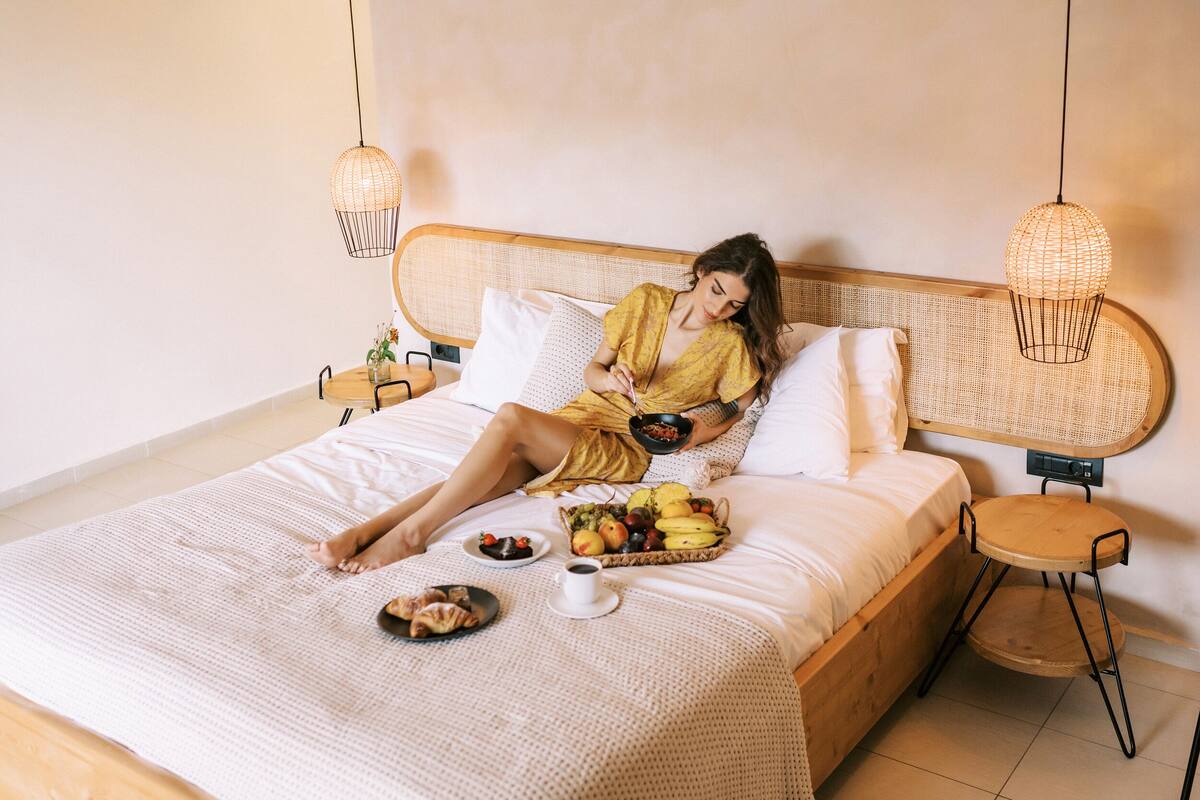 Una cama con cabecera de mimbre, una mesita de noche con cesta de frutas y un taburete pequeño.