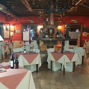 Un restaurante con varias mesas, cada una cubierta con un mantel rojo y blanco. Hay botellas de vino en las mesas, y las paredes están decoradas con diversos adornos.