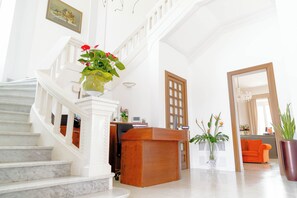 A spacious lobby with a marble staircase, a wooden reception desk, and a large potted plant. - Villa Mosca (Alghero)