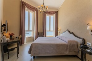 A hotel room with a large bed, a desk with a chair, a chandelier, and a view of the sea through the windows. - Villa Mosca (Alghero)