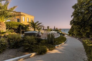 A pathway leading to a building with a pool and outdoor seating area, surrounded by tropical plants and a view of the sea.