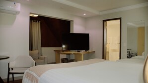 Une chambre d’hôtel avec un grand lit, un téléviseur posé sur un meuble en bois, un bureau avec une chaise et une salle de bain visible à travers une porte ouverte.